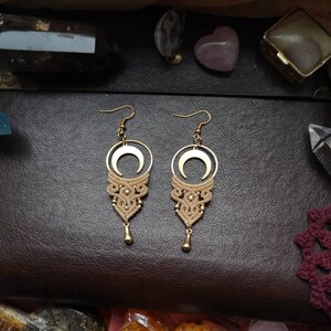 May include: A pair of gold crescent moon earrings with a beige macrame design hanging from the bottom. The earrings are on a brown leather surface with crystals and other jewelry.