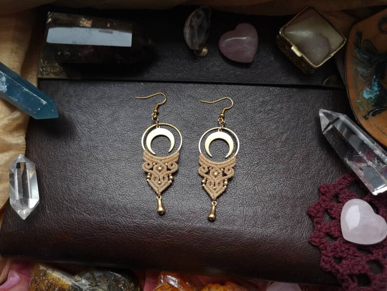 May include: A pair of gold crescent moon earrings with a beige macrame design hanging from the bottom. The earrings are on a brown leather surface with crystals and other jewelry.