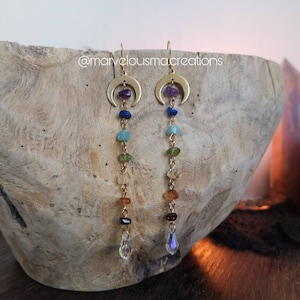 May include: Gold crescent moon earrings with a dangling chain of colorful gemstones. The gemstones are in the order of the chakras: purple, blue, green, yellow, orange, and red. The earrings are finished with a clear crystal drop.