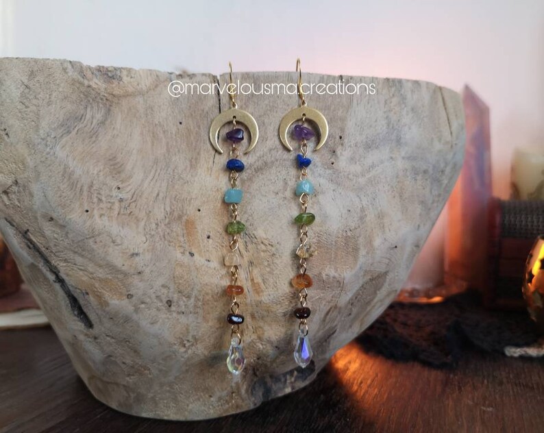 May include: Gold crescent moon earrings with a dangling chain of colorful gemstones. The gemstones are in the order of the chakras: purple, blue, green, yellow, orange, and red. The earrings are finished with a clear crystal drop.