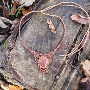 Puede incluir: Un collar de macram&eacute; marr&oacute;n con un colgante de piedra de sol roja. El collar est&aacute; hecho con un cord&oacute;n anudado y tiene un dise&ntilde;o simple. El colgante es una piedra redonda y plana de color marr&oacute;n rojizo.