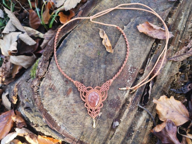 Puede incluir: Un collar de macram&eacute; marr&oacute;n con un colgante de piedra de sol roja. El collar est&aacute; hecho con un cord&oacute;n anudado y tiene un dise&ntilde;o simple. El colgante es una piedra redonda y plana de color marr&oacute;n rojizo.