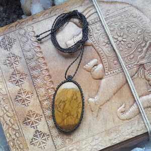 May include: A brown and gold oval stone pendant on a black cord necklace. The pendant is on a brown leather surface with an embossed elephant design.
