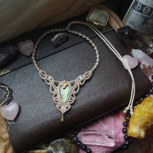Puede incluir: Un collar de macram&eacute; con un colgante de piedra iridiscente verde y dorada. El collar est&aacute; hecho con cord&oacute;n beige y tiene un dise&ntilde;o delicado e intrincado. El colgante est&aacute; rodeado de peque&ntilde;as cuentas blancas.