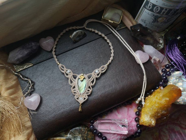 Puede incluir: Un collar de macram&eacute; con un colgante de piedra iridiscente verde y dorada. El collar est&aacute; hecho con cord&oacute;n beige y tiene un dise&ntilde;o delicado e intrincado. El colgante est&aacute; rodeado de peque&ntilde;as cuentas blancas.