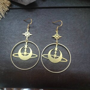 May include: A pair of gold-toned earrings featuring a crescent moon and star design. The earrings are made of metal and have a hoop shape.