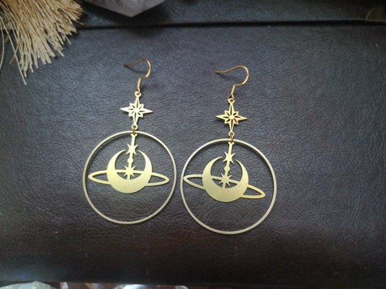 May include: A pair of gold-toned earrings featuring a crescent moon and star design. The earrings are made of metal and have a hoop shape.