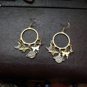 May include: A pair of gold hoop earrings with dangling charms. The charms include a crescent moon, a bee, a butterfly, a star, and a planet with a ring around it. Each charm is decorated with sparkling rhinestones.