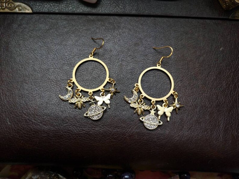 May include: A pair of gold hoop earrings with dangling charms. The charms include a crescent moon, a bee, a butterfly, a star, and a planet with a ring around it. Each charm is decorated with sparkling rhinestones.