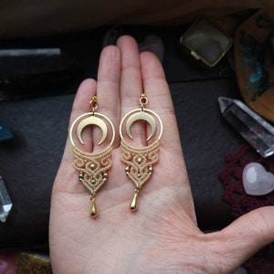May include: A pair of gold crescent moon earrings with a delicate macrame design and a small gold teardrop charm hanging from each earring.