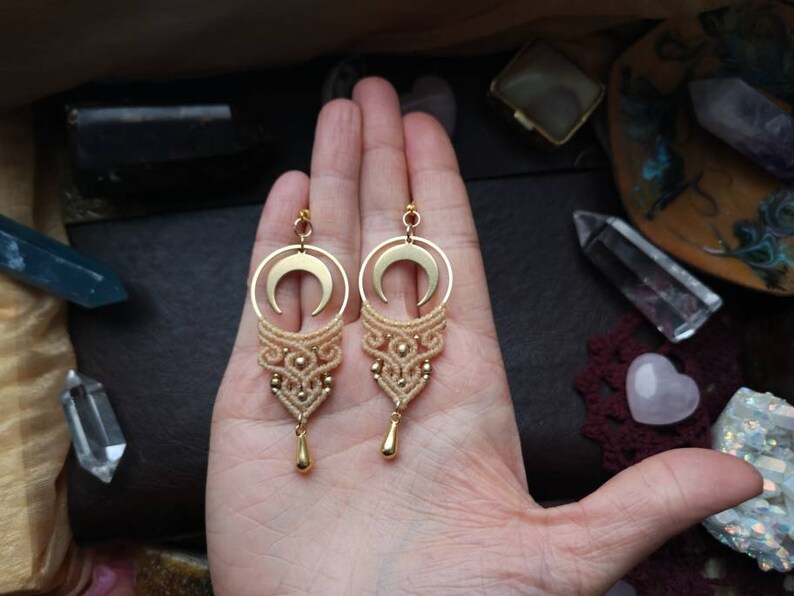 May include: A pair of gold crescent moon earrings with a delicate macrame design and a small gold teardrop charm hanging from each earring.