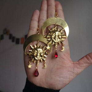 May include: Gold tone earrings with a crescent moon and sun design. Each earring features a sun with a face, a crescent moon, and dangling red crystal drops.