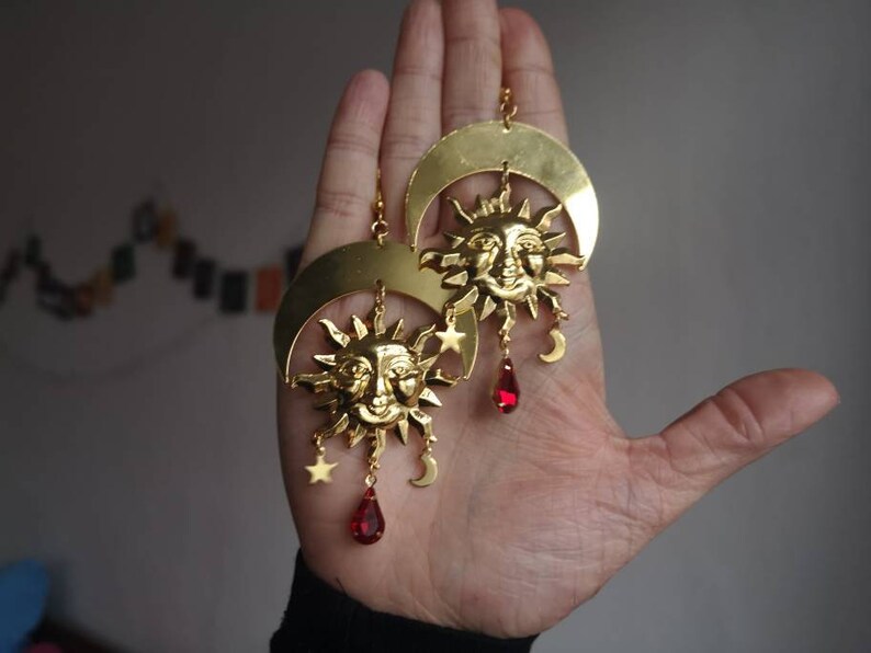 May include: Gold tone earrings with a crescent moon and sun design. Each earring features a sun with a face, a crescent moon, and dangling red crystal drops.
