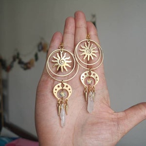 May include: Gold tone earrings with a sun and moon design. The earrings feature a circle with a sun design and a crescent moon with stars. The earrings are accented with clear crystal beads and gold tone spikes.