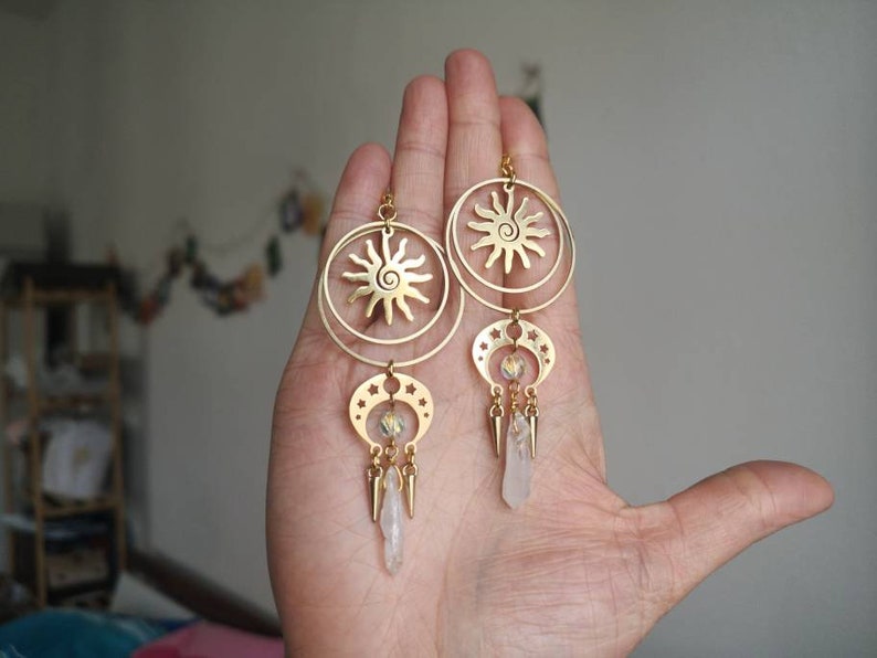 May include: Gold tone earrings with a sun and moon design. The earrings feature a circle with a sun design and a crescent moon with stars. The earrings are accented with clear crystal beads and gold tone spikes.