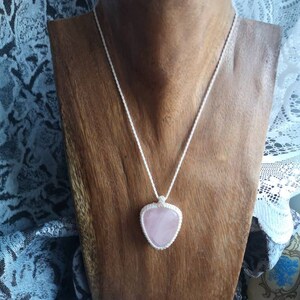May include: A pink rose quartz heart pendant necklace on a white cord. The necklace is displayed on a wooden mannequin.