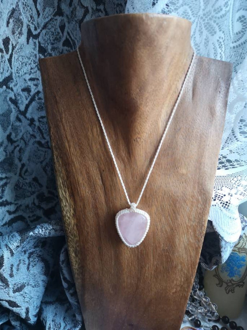 May include: A pink rose quartz heart pendant necklace on a white cord. The necklace is displayed on a wooden mannequin.