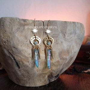 May include: Gold earrings with a sun and moon design, featuring iridescent blue crystal accents. The earrings are hanging from a wooden surface.