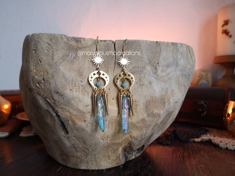 May include: Gold earrings with a sun and moon design, featuring iridescent blue crystal accents. The earrings are hanging from a wooden surface.