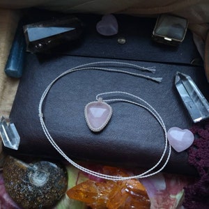 May include: A pink rose quartz pendant necklace with a white cord. The necklace is lying on a brown leather surface with other crystals and stones.