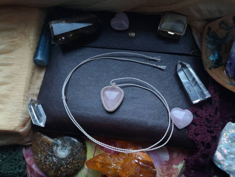 May include: A pink rose quartz pendant necklace with a white cord. The necklace is lying on a brown leather surface with other crystals and stones.