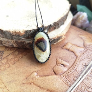 May include: A brown and white agate pendant with black inclusions, hanging from a black cord necklace. The pendant is oval shaped and has a natural, organic look.