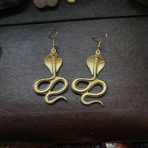 May include: A pair of gold-toned earrings shaped like a cobra snake. The earrings are dangling and have a detailed design.