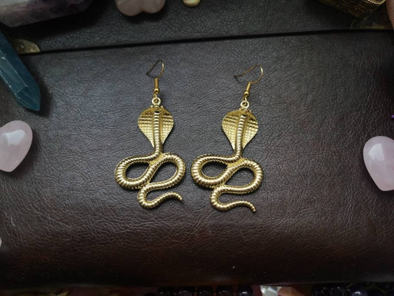 May include: A pair of gold-toned earrings shaped like a cobra snake. The earrings are dangling and have a detailed design.