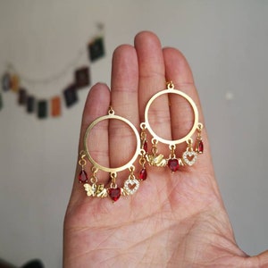 May include: Gold hoop earrings with dangling charms. The charms are red heart-shaped stones with gold accents and small gold wings.