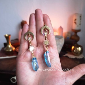 K&ouml;nnte beinhalten: Goldene Ohrringe mit einem Halbmond-Anh&auml;nger, einem Sonnen-Anh&auml;nger und einem blauen Kristall-Anh&auml;nger. Die Ohrringe h&auml;ngen an der Hand einer Person.