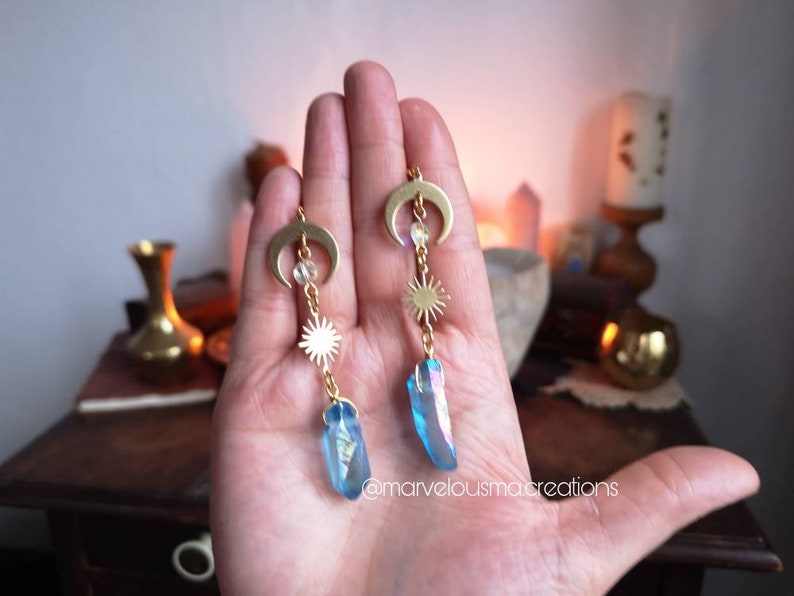K&ouml;nnte beinhalten: Goldene Ohrringe mit einem Halbmond-Anh&auml;nger, einem Sonnen-Anh&auml;nger und einem blauen Kristall-Anh&auml;nger. Die Ohrringe h&auml;ngen an der Hand einer Person.