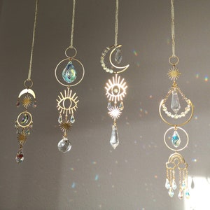 Könnte beinhalten: Fünf goldfarbene Kristall-Sonnenfänger hängen von der Decke. Jeder Sonnenfänger hat ein anderes Design, darunter einen Halbmond, ein Auge und einen Regenbogen. Die Sonnenfänger sind aus Metall und Glas und haben verschiedene Kristallformen.
