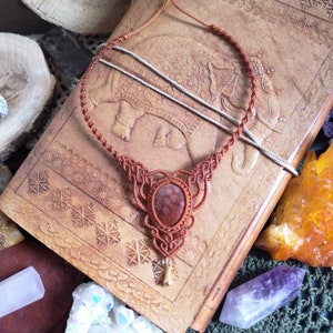 Puede incluir: Un collar de cord&oacute;n de cuero marr&oacute;n con un dise&ntilde;o de macram&eacute; y un colgante de piedra marr&oacute;n rojizo.