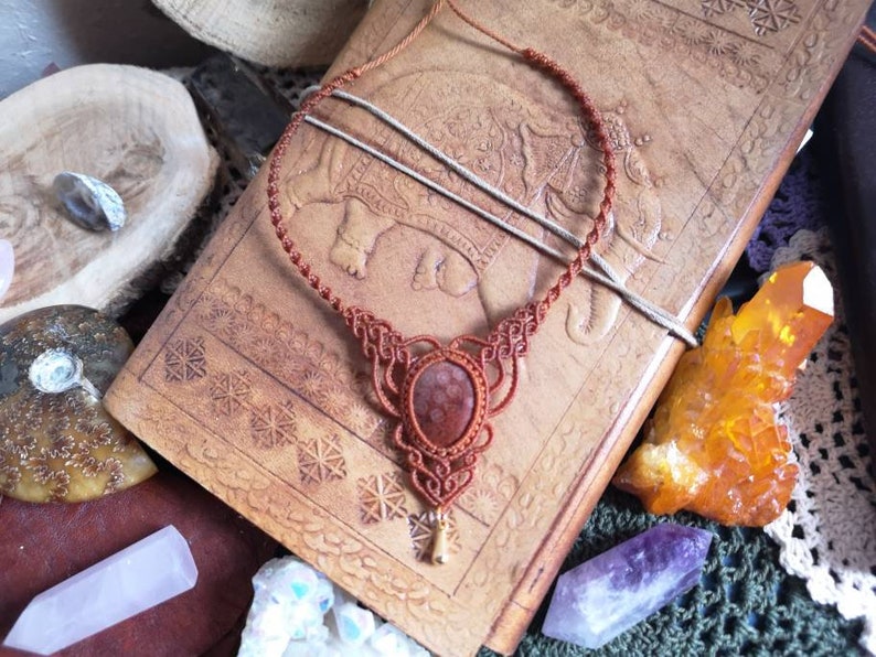Puede incluir: Un collar de cord&oacute;n de cuero marr&oacute;n con un dise&ntilde;o de macram&eacute; y un colgante de piedra marr&oacute;n rojizo.