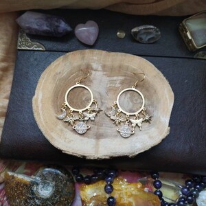 May include: Gold hoop earrings with dangling charms, including a crescent moon, stars, butterflies, and a small globe. The charms are adorned with sparkling rhinestones.