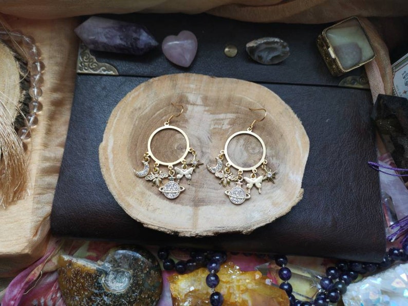 May include: Gold hoop earrings with dangling charms, including a crescent moon, stars, butterflies, and a small globe. The charms are adorned with sparkling rhinestones.