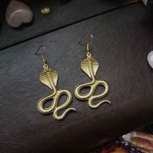 May include: A pair of gold-toned snake earrings. Each earring features a detailed cobra design with a raised, textured pattern.