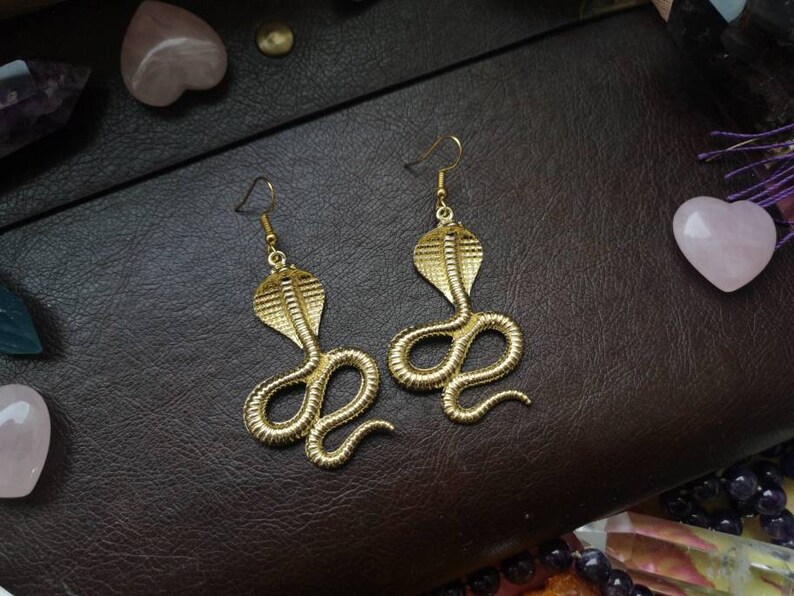 May include: A pair of gold-toned snake earrings. Each earring features a detailed cobra design with a raised, textured pattern.