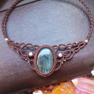May include: A brown cord necklace with a large oval-shaped blue-green gemstone pendant. The pendant is surrounded by a intricate knotwork design with small white beads.