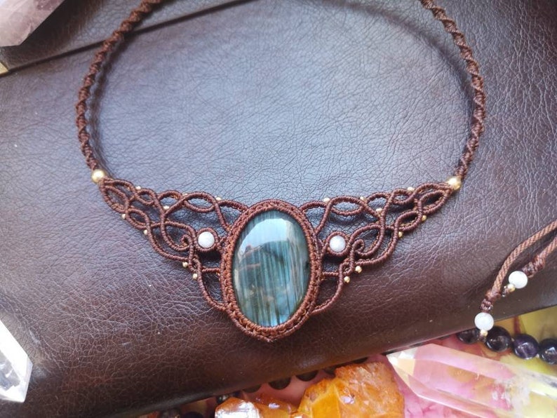 May include: A brown cord necklace with a large oval-shaped blue-green gemstone pendant. The pendant is surrounded by a intricate knotwork design with small white beads.