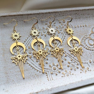 May include: Gold tone earrings with a crescent moon, star, and sword design. The earrings are dangling and have a delicate, intricate design.