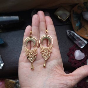 May include: A pair of gold crescent moon earrings with macrame detailing in a light beige color. The earrings are hanging from a hand.