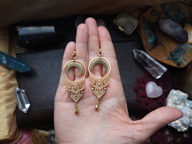 May include: A pair of gold crescent moon earrings with macrame detailing in a light beige color. The earrings are hanging from a hand.
