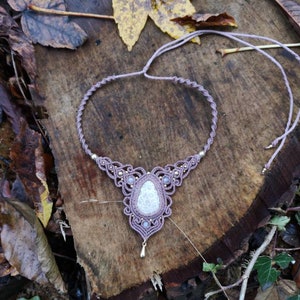May include: A delicate, mauve macrame necklace with a white gemstone pendant. The necklace is made with a knotted cord and features small gold beads.