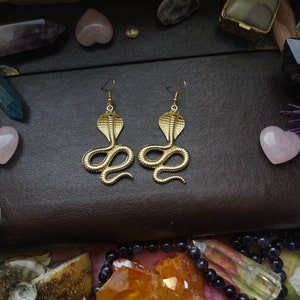 May include: A pair of gold-toned earrings shaped like a cobra snake. The earrings are on a brown leather surface with various crystals and stones.