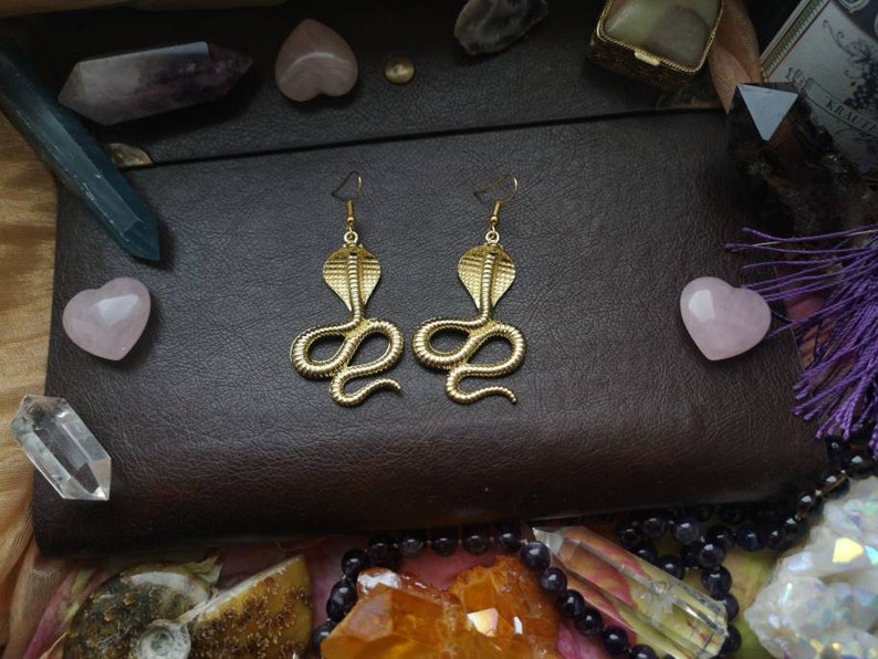 May include: A pair of gold-toned earrings shaped like a cobra snake. The earrings are on a brown leather surface with various crystals and stones.