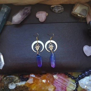 Puede incluir: Un par de pendientes de oro con un dise&ntilde;o de luna creciente y un amuleto de mano de hamsa. Los pendientes est&aacute;n adornados con gotas de cristal de color p&uacute;rpura y azul degradado.