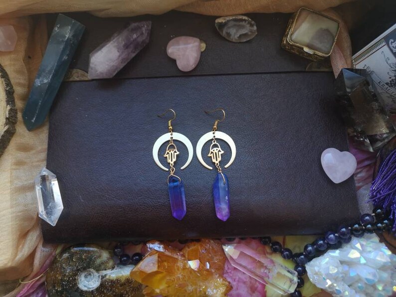 Puede incluir: Un par de pendientes de oro con un dise&ntilde;o de luna creciente y un amuleto de mano de hamsa. Los pendientes est&aacute;n adornados con gotas de cristal de color p&uacute;rpura y azul degradado.