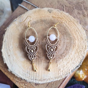May include: A pair of handmade earrings with a delicate, intricate design. The earrings feature a light pink gemstone bead and a gold-colored metal accent. The earrings are suspended from a gold-colored hook.