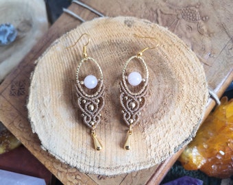 May include: A pair of handmade earrings with a delicate, intricate design. The earrings feature a light pink gemstone bead and a gold-colored metal accent. The earrings are suspended from a gold-colored hook.